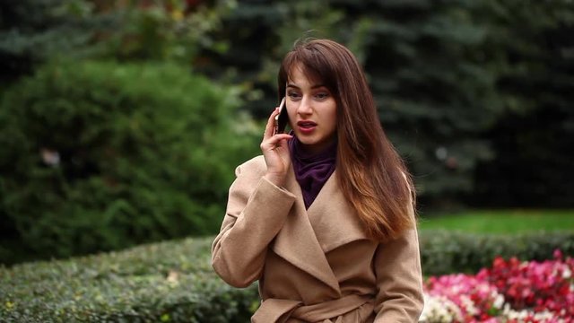 Young beautiful brunette woman in beige coat sitting on a bench in the autumn park