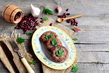 Cutlets cooked from mashed red beans. Vegetarian cutlets, scattered raw red beans, garlic, fresh parsley, fork, knife, small wooden spoon and decorative barrel on old wooden background. Vintage style