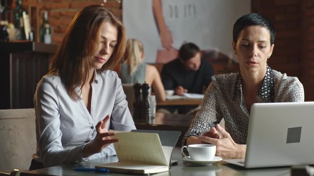 Two businesswomen sitting at cafe table, using laptop and talking