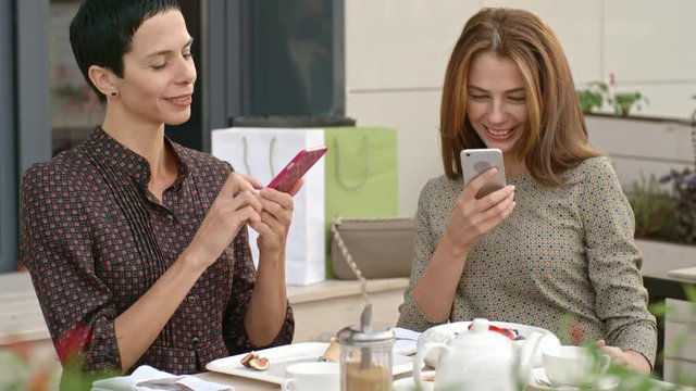 Two women sitting in the street cafe and taking pictures of food with smart phones 