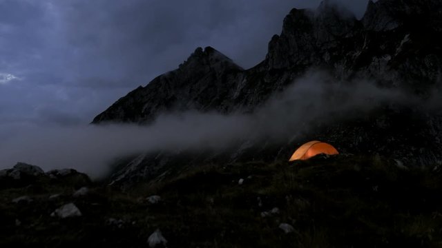 Camping in the Night. Reveal of a beautifully lit tent with high mountain tops in background.
