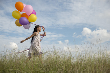 Cute little girl holding colorful balloons, running in the meadow against blue sky and clouds.