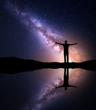 Milky Way. Night starry and silhouette of a standing alone man with with raised up arms on the mountain near the lake with sky reflection in water. Milky way and man on the hill. Galaxy. Universe