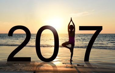 Happy new year card 2017. Silhouette of A girl doing Yoga vrikshasana tree pose on tropical  beach with sunset sky background, watching the sunset, standing as a part of the Number 2017 sign.