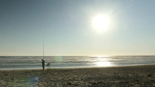 Elderly fisherman fidgeting with his rod during golden hour at a deserted beach.
