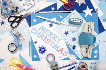 Arts and craft supplies for Christmas. Blue color paper, pencils, different washi tapes, craft scissors, wrapping paper rolls, festive Xmas supplies for decoration. Merry Christmas paper sign.