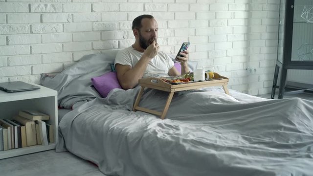 Young man using smartphone during breakfast on bed, 4K
