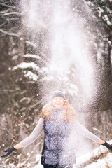 Fototapeta na wymiar Winter fun in old wood. Girl in winter clothes throwing snow up in air. Young attractive woman enjoys snowy winter. Portrait of beautiful woman with long ginger hair over trees background.