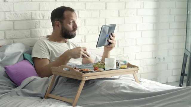 Young man watching movie on tablet and eating breakfast while lying on bed at home
