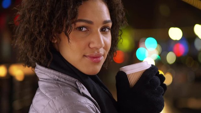 Closeup of black woman holding cup, looking at camera in with bokeh lights.
