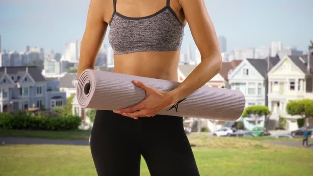 Active black woman holding rolled yoga mat on grass.