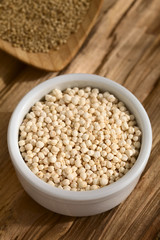 Popped white quinoa (lat. Chenopodium quinoa) cereal in small bowl, raw quinoa grains in the back, photographed on wood with natural light (Selective Focus, Focus one third into the quinoa cereal)