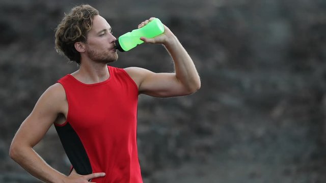 Running man - portrait of male runner resting drinking water from water bottle after cross country run on trail. Fit handsome athlete taking break standing sweating after workout outside in nature.