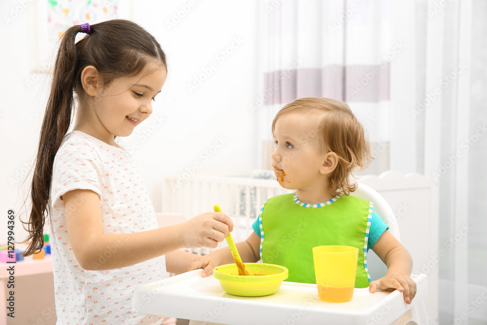 Poster Cute girl feeding little sister in high chair at baby room