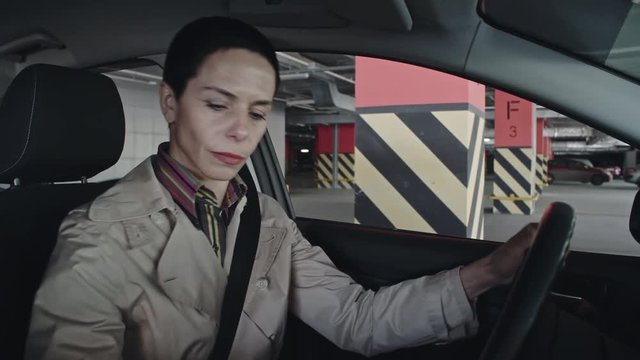 Shot from inside of automobile cabin of serious female driver with short hair reversing car into parking slot, then leaving