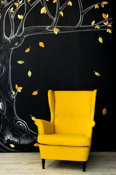 A comfortable bright yellow armchair opposite a black wall with a painted autumn tree with falling yellow leaves.