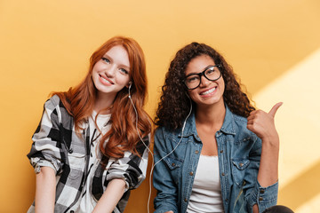 Fototapeta premium Two happy lovely young women with earphones showing thumbs up