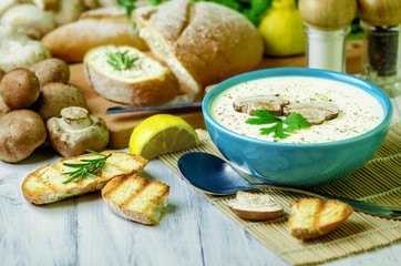 Delicious creamy mushroom soup in blue bowl placed with fresh ciabatta bread,lemon and organic mushroom on white wooden background..