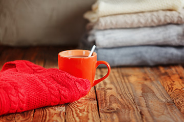 Mug with cacao or tea and red sweater on wooden table  stack sweaters background.