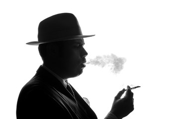 Silhouette of private detective lights cigarette. Agent looks like Al Capone stay side to camera. Police criminal scene in black and white. Gangster studio shot