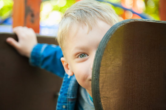 Close up of half of smiling beautiful child face hiding behind wooden element of children slide at playground. Portrait of cute funny little boy dressed in blue denim clothes. Horizontal color image.