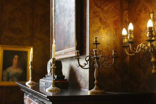 Two golden coloured candleholders with lit candles in a classical fireplace with a painting of a woman in the background.