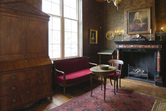 A sitting corner with a red bench, small table, chair, large wooden cabinet and a dark fireplace near a window in a traditional Dutch interior.