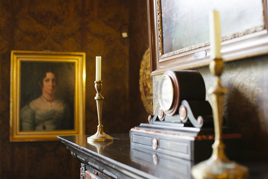 Two golden coloured candleholders in a classical fireplace with a painting of a woman in the background.