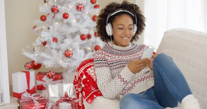 Attractive woman listening to music on her mobile phone at Christmas as she relaxes on a sofa in front of the tree with a happy smile.