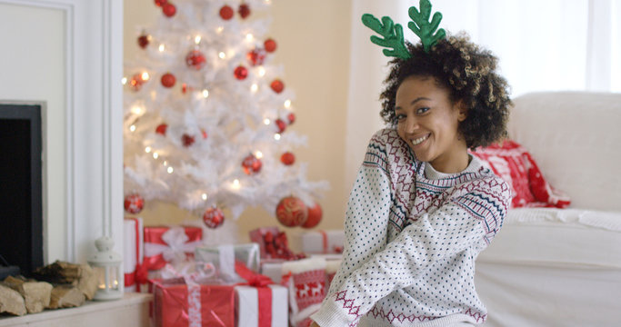 Cute young woman posing in green reindeer antlers in a festive red and white living room in front of the Christmas tree smiling at the camera.