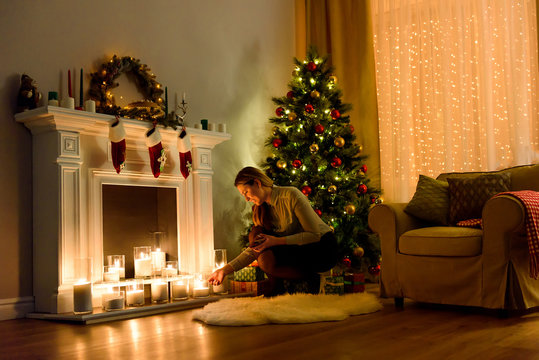 A nice woman in a cosily lighted christmas decorated room fixing candles on a fireplace. Christmas Room Interior Design. Copyspace. Xmas tree decorated by lights, candles and indoors fireplace.