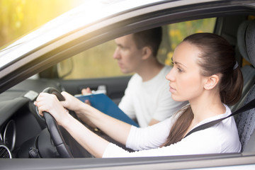 Driving test. Young serious woman driving car feeling inexperienced, looking nervous at the road traffic for information to make appropriate decisions. Man is an instructor, controlling and checking