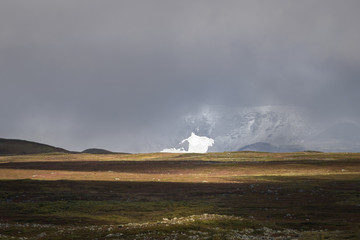 Fototapeta premium Sunlight giving golden color to the autumn colored landscape with glacier covered in the mist and clouds