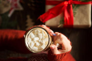 Closeup on hot chocolate with marshmallows in hand of happy woman against the background of a table with Christmas gifts

