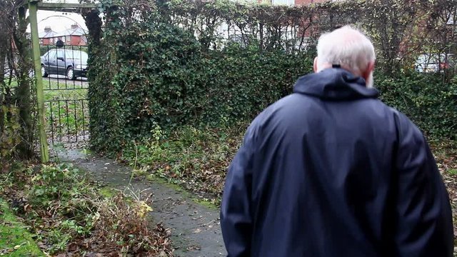 Middle aged man in cagoule on a wet autumn day, purposefully striding up a garden path away from the house and out of the garden gate. The garden is a little unkempt and covered with fallen leaves.