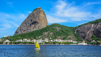 Yellow small sailing yacht and classic daytime scenic profile view of Sugarloaf Mountain, Pao de Acucar, standing above Botafogo Bay in Rio de Janeiro, Brazil