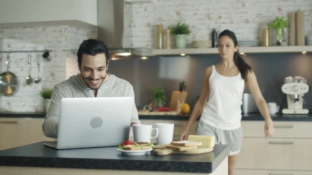 In the Kitchen Young Man Sits at the Table Laptop before Him, He Calls His Gorgeous Girfriend and Shows Her Something on the Screen. They Both Laugh. Shot on RED Cinema Camera in 4K (UHD).