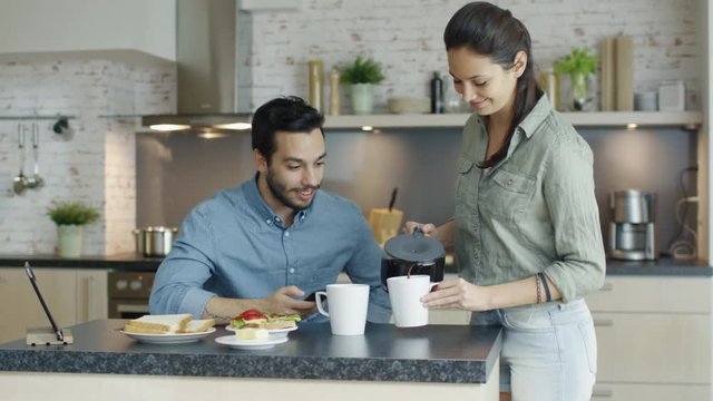 In the Kitchen Handsome Young Man Sits at the Table and Uses Smartphone, Beautiful Girl Brings Coffee Pot with Fresh Coffee.  Shot on RED Cinema Camera in 4K (UHD).