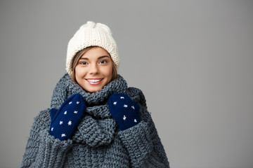 Young beautiful fair-haired girl in knited hat sweater and mittens smiling looking at camera over grey background. 