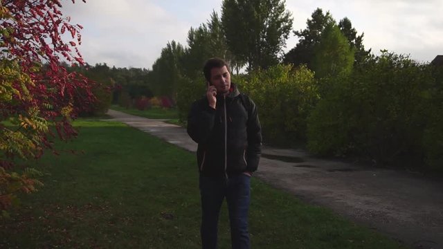 Young man in black jacket walking and talking on the phone in the autumn park slow motion HD