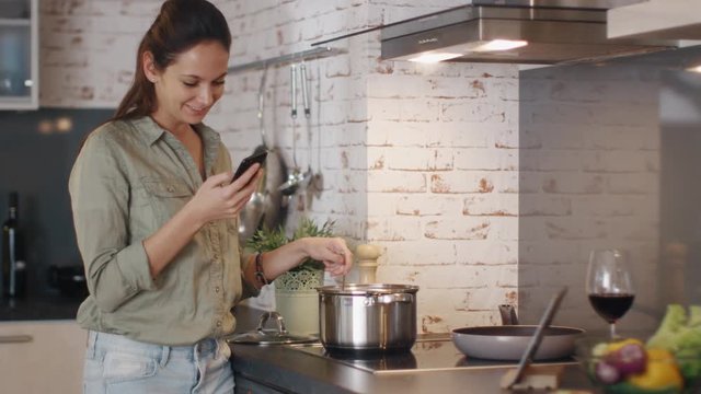 Young Woman Stirs Food in Pan While Holding Her Smartphone and Smiling. Shot on RED Cinema Camera in 4K (UHD).
