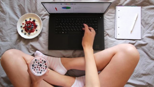 Overhead shot of woman in bed, feet , laptop, yogurt and notebook on the gray blanket