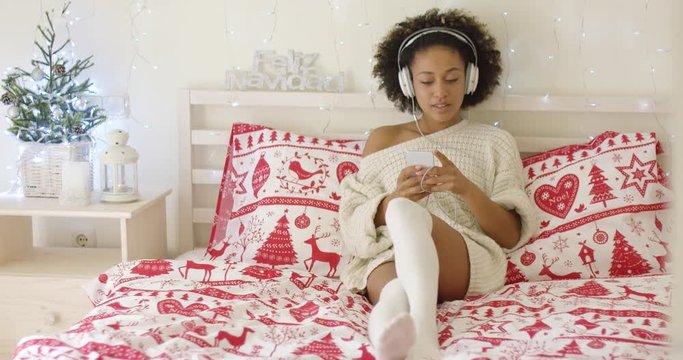 Beautiful young adult woman in long sweater and stocking resting on bed while listening to music on her mp3 player