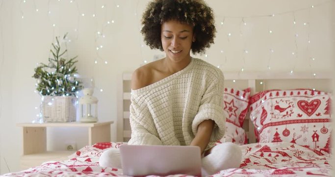 Single happy young adult woman in oversized sweater on bed using laptop computer. Includes Christmas theme blankets and lights.