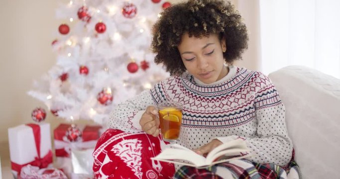 Young woman relaxing on a sofa with a cup of tea reading a book in front of a Christmas tree