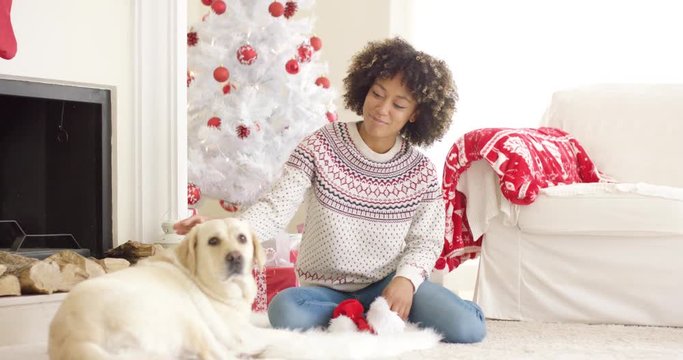 Pretty smiling young woman stroking her golden retriever dog as they relax together on the floor in front of a Christmas tree and hearth