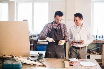 Happy male carpenter showing something to coworker at his notebook papers in workshop