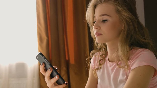 Blond woman sitting on the bed, holding her phone in hands and chatting to hear friends in a pink pajama.