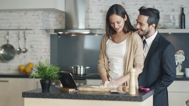 Young Girl Prepares Flour for Cooking. For Recipe Reference Tablet Stands on the Kitchen Table. Handsome Businessman Boyfriend comes to Her and Hugs. Both Smile. Shot on RED Cinema Camera in 4K (UHD).