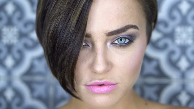 Closeup face of beautiful girl with silver smoky eye makeup and pink lips opening eyes and looking into the camera over gray vintage wall background - video in slow motion